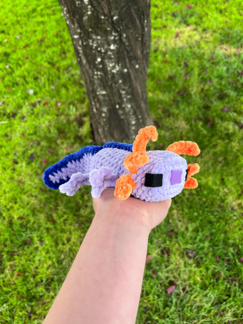 Op de afbeelding: Een handgemaakte gehaakte axolotl-knuffel in paars, blauw en oranje. Het pluche speelgoed heeft een gedetailleerd ontwerp met een getextureerd lichaam, kleine oranje kieuwen en een vierkante kop. De axolotl wordt vastgehouden tegen een achtergrond van groen gras en een boomstam.