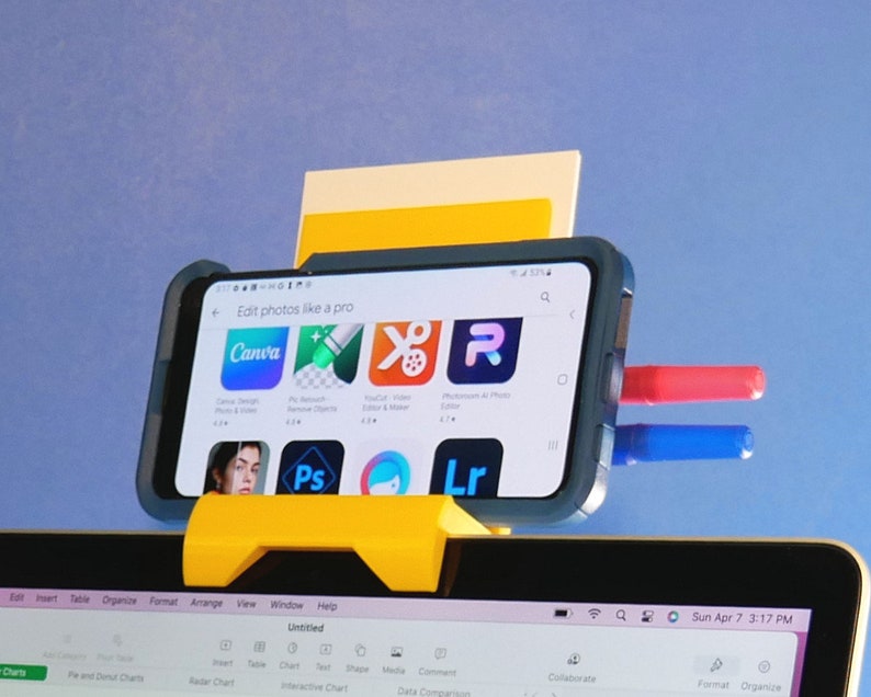 May include: A yellow phone holder with a smartphone displaying app icons, including Canva. The phone is propped up with red and blue markers. The screen reads "Edit photos like a pro". A laptop is visible below.
