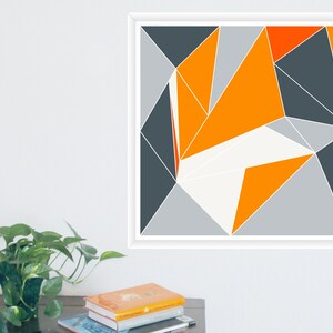 May include: Geometric abstract art print in a white frame. The artwork features a design of triangles in shades of orange, grey, and white. The artwork is displayed on a white wall.