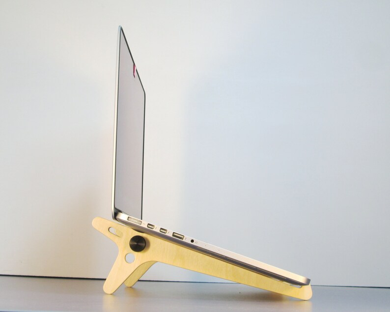 May include: A light-colored wooden laptop stand with a minimalist design. The stand is angled, supporting a silver laptop. It features a circular knob and a three-legged base. The laptop is open, displaying a bright screen.