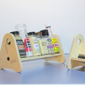 May include: Two wooden spice racks with a light finish. Each rack has a triangular side with a circular hole and holds several spice jars and bottles. The jars contain various spices, including cumin, sea salt, and garlic.