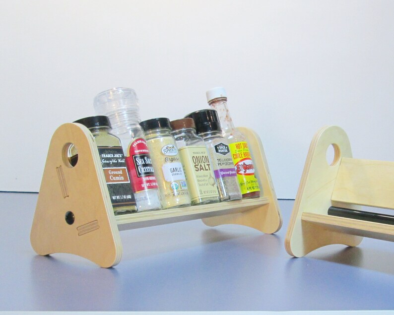 May include: Two wooden spice racks with a light finish. Each rack has a triangular side with a circular hole and holds several spice jars and bottles. The jars contain various spices, including cumin, sea salt, and garlic.