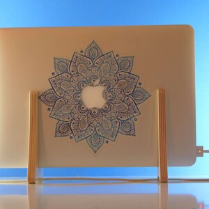 May include: A silver laptop with a blue and white mandala design sticker is supported by a wooden stand. The laptop is connected to a charger. The background is a blue gradient.