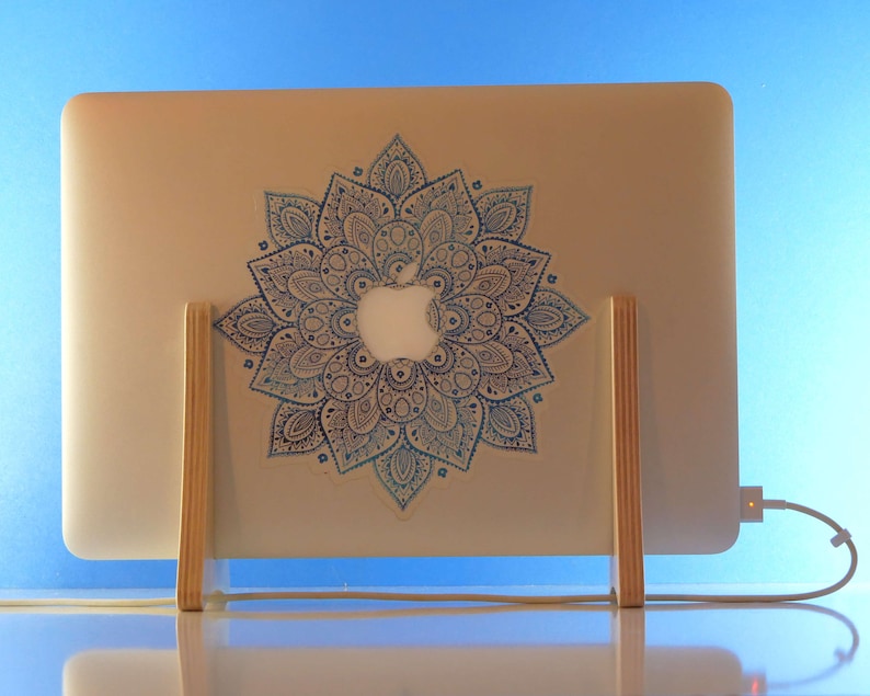 May include: A silver laptop with a blue and white mandala design sticker is supported by a wooden stand. The laptop is connected to a charger. The background is a blue gradient.
