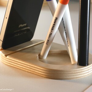 May include: A wooden desk organizer with a slot for a smartphone and holes for pens and markers. The organizer holds a black iPhone, two white Paper Mate Flair pens, and a black pen. The organizer is made of layered wood.