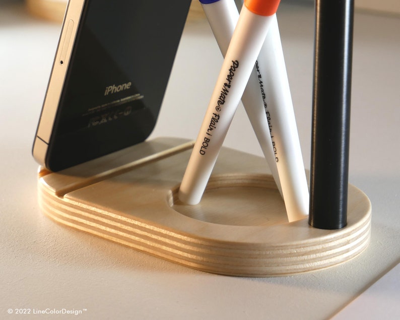 May include: A wooden desk organizer with a slot for a smartphone and holes for pens and markers. The organizer holds a black iPhone, two white Paper Mate Flair pens, and a black pen. The organizer is made of layered wood.