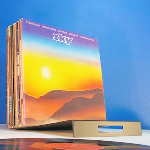 May include: A stack of vinyl records in a gray metal holder. The album cover features a sunset over mountains with the word "Sky" in white. The background is a gradient of yellow, orange, and blue.