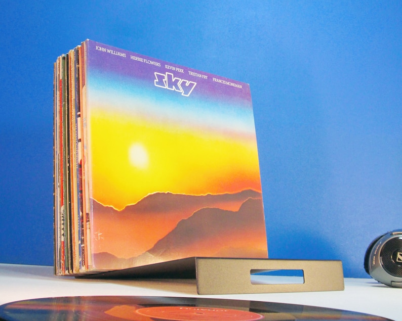 May include: A stack of vinyl records in a gray metal holder. The album cover features a sunset over mountains with the word "Sky" in white. The background is a gradient of yellow, orange, and blue.