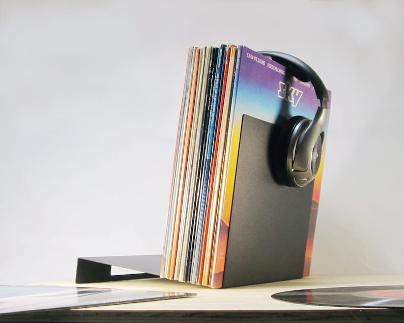 May include: A black metal record holder with a stack of vinyl records. A pair of silver headphones rests on the holder. The record covers display various colors and designs, with one labeled "Sky".