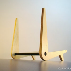 May include: A wooden stand with a natural finish. The stand has a unique design with two upright supports connected by a horizontal bar. The supports are angled and have a curved top. A small foot provides stability. The stand is likely for a tablet or book.