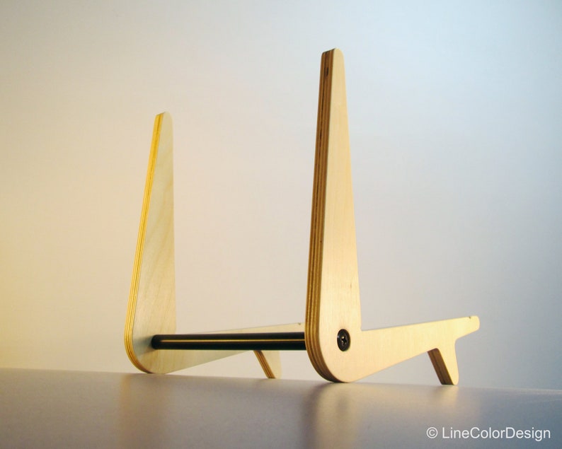May include: A wooden stand with a natural finish. The stand has a unique design with two upright supports connected by a horizontal bar. The supports are angled and have a curved top. A small foot provides stability. The stand is likely for a tablet or book.
