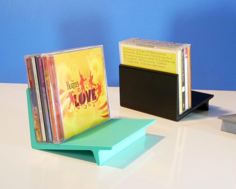 May include: Two CD holders, one teal and one black, each holding a stack of CDs. The teal holder has a slanted design, and the black holder is rectangular. The Beatles "LOVE" album is visible in the teal holder.