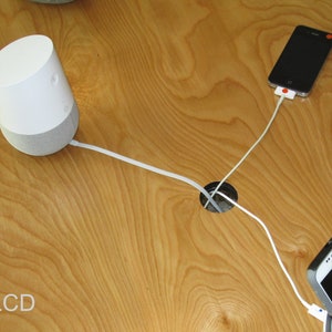 May include: A white and gray smart speaker with a white cable connected to a wooden surface. Two smartphones are also connected to the surface with white cables. The text "LCD" is visible in the lower left corner.