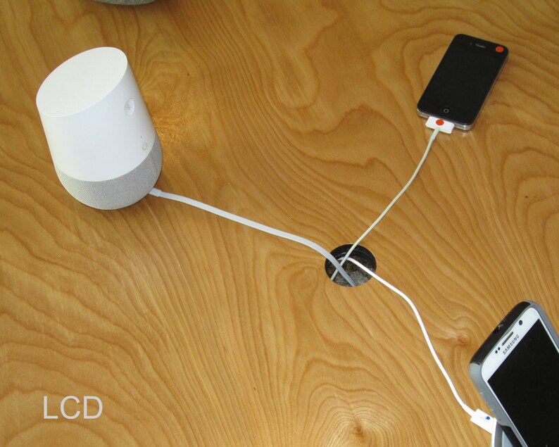 May include: A white and gray smart speaker with a white cable connected to a wooden surface. Two smartphones are also connected to the surface with white cables. The text "LCD" is visible in the lower left corner.