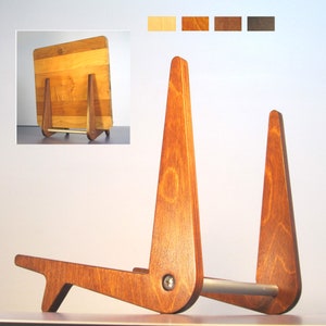 May include: A wooden book stand with a warm, reddish-brown finish. The stand features a unique, angled design with a square back and a horizontal bar for support. The wood grain is visible, adding to its natural aesthetic. The stand is designed to hold a book or tablet.
