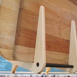 May include: Wooden cutting boards in a light wood tone are displayed in a light wood stand. The stand has a minimalist design with a black bar at the bottom. The cutting boards are various sizes and shapes.