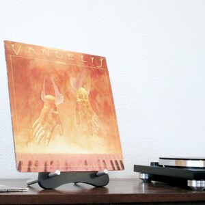 May include: A vinyl record album titled "Vangelis" displayed on a black stand. The album cover features a warm-toned illustration of two hands with wings. A turntable is visible in the background, suggesting a music theme.