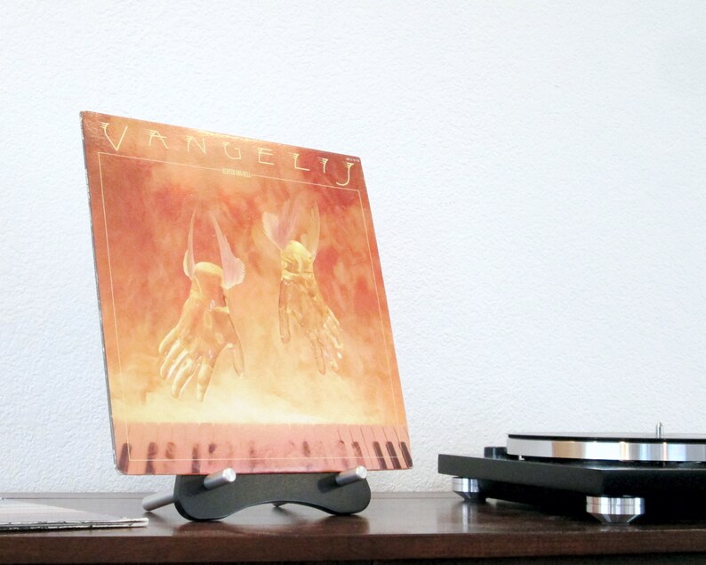 May include: A vinyl record album titled "Vangelis" displayed on a black stand. The album cover features a warm-toned illustration of two hands with wings. A turntable is visible in the background, suggesting a music theme.