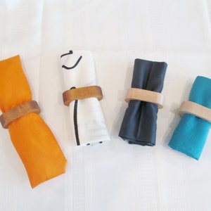 May include: Four cloth napkins in orange, white, navy blue, and teal, each rolled and held by a wooden napkin ring. The napkins are arranged on a white surface, showcasing a simple yet elegant table setting.