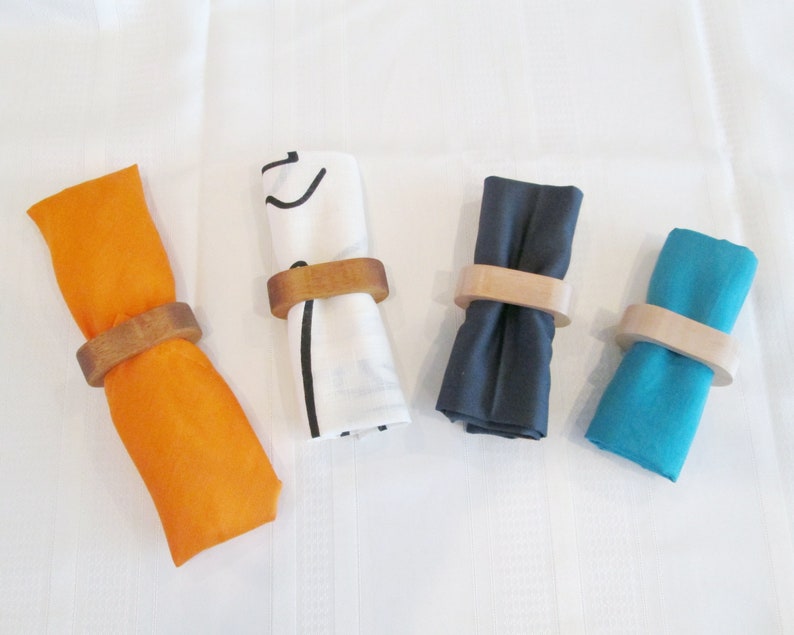 May include: Four cloth napkins in orange, white, navy blue, and teal, each rolled and held by a wooden napkin ring. The napkins are arranged on a white surface, showcasing a simple yet elegant table setting.