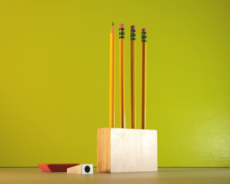 May include: A still life composition featuring four yellow pencils with pink erasers in a wooden pencil holder. A red eraser and a pencil sharpener are also present. The background is a solid lime green.