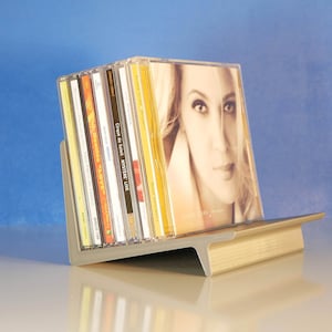 May include: A gray and gold CD holder displaying several CDs. The CDs are in clear plastic cases, with album art visible. The holder is made of metal and has a modern design, with a slanted back and a flat base. The background is a gradient of blue.