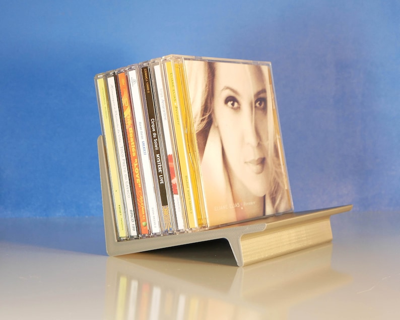 May include: A gray and gold CD holder displaying several CDs. The CDs are in clear plastic cases, with album art visible. The holder is made of metal and has a modern design, with a slanted back and a flat base. The background is a gradient of blue.