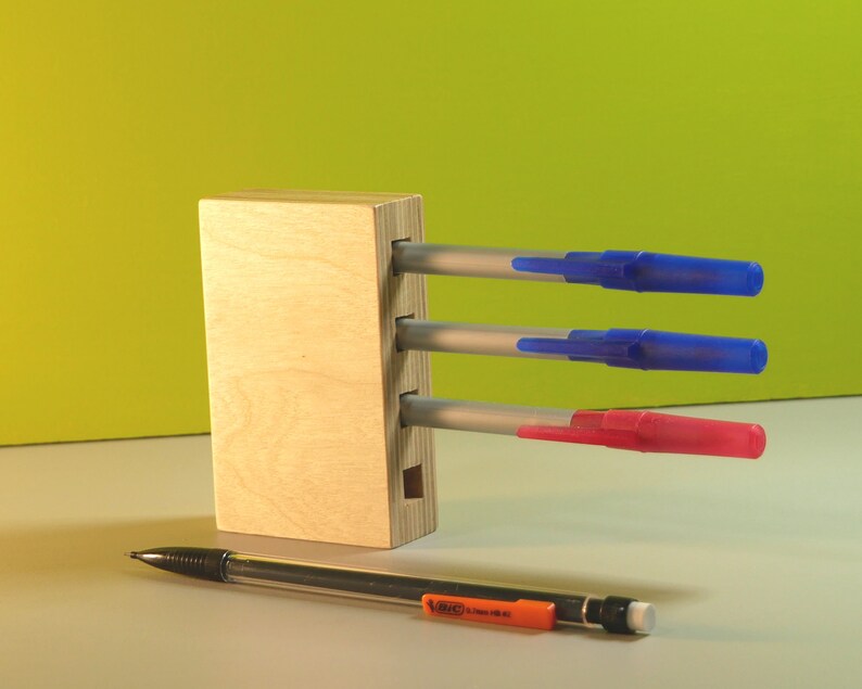May include: A wooden pen holder with three pens and a pencil. The pens have blue and red caps. The pencil is black with an orange eraser holder. The background is a gradient of green and yellow.