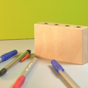 May include: A light-colored wooden desk organizer with five holes on top, holding several pens. The pens are blue, pink, and silver. The background is a light green color.