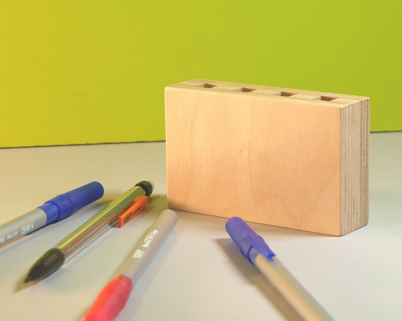 May include: A light-colored wooden desk organizer with five holes on top, holding several pens. The pens are blue, pink, and silver. The background is a light green color.