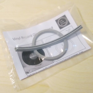 May include: A vinyl record display stand kit in a clear plastic bag. The kit includes a gray curved support, two gray rings, and a black disc. The packaging includes a printed sheet with the text "Vinyl Record Display".