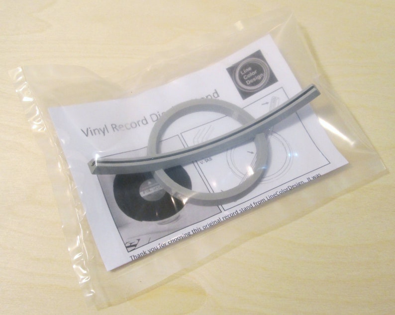 May include: A vinyl record display stand kit in a clear plastic bag. The kit includes a gray curved support, two gray rings, and a black disc. The packaging includes a printed sheet with the text "Vinyl Record Display".