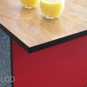 May include: A modern coffee table with a light wood grain top and a red base. The top has a black edge. Two glasses of orange juice sit on the table. The table is a stylish addition to any living room.