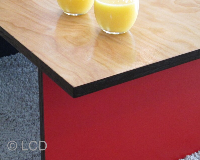 May include: A modern coffee table with a light wood grain top and a red base. The top has a black edge. Two glasses of orange juice sit on the table. The table is a stylish addition to any living room.