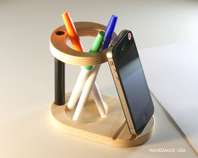May include: A wooden desk organizer with a circular top and base, holding pens and a smartphone. The organizer has a natural wood finish with black accents. Pens are orange, green, and blue. The phone is black and gold. Text reads "HANDMADE USA".