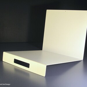 May include: A white, L-shaped metal shelf with a rectangular cutout. The shelf has a flat base and a vertical back panel. The design is minimalist and modern, with a clean, smooth surface. The background is dark.