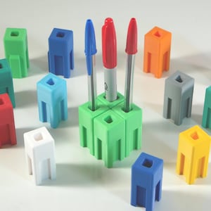May include: A collection of colorful pen holders in various shades, including green, blue, red, and yellow. The pen holders are cube-shaped with a square opening at the top and three legs. Pens and markers are placed in the green pen holder. The pen holders are arranged on a white surface.