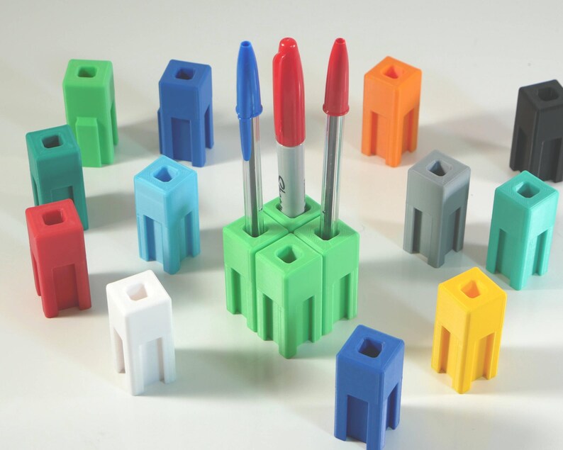 May include: A collection of colorful pen holders in various shades, including green, blue, red, and yellow. The pen holders are cube-shaped with a square opening at the top and three legs. Pens and markers are placed in the green pen holder. The pen holders are arranged on a white surface.