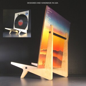 May include: A wooden record album display stand holding a vinyl record. The stand is made of light-colored wood and has a minimalist design. The album cover features a sunset landscape. Text on the top reads: DESIGNED AND HANDMADE IN USA.