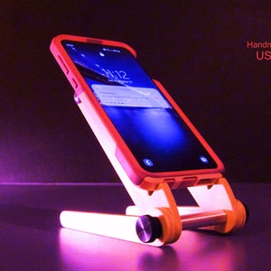 May include: A wooden phone stand with a red-orange phone case. The stand is made of light-colored wood and has a minimalist design. The phone displays the time and date. Text on the image reads "Handmade USA".