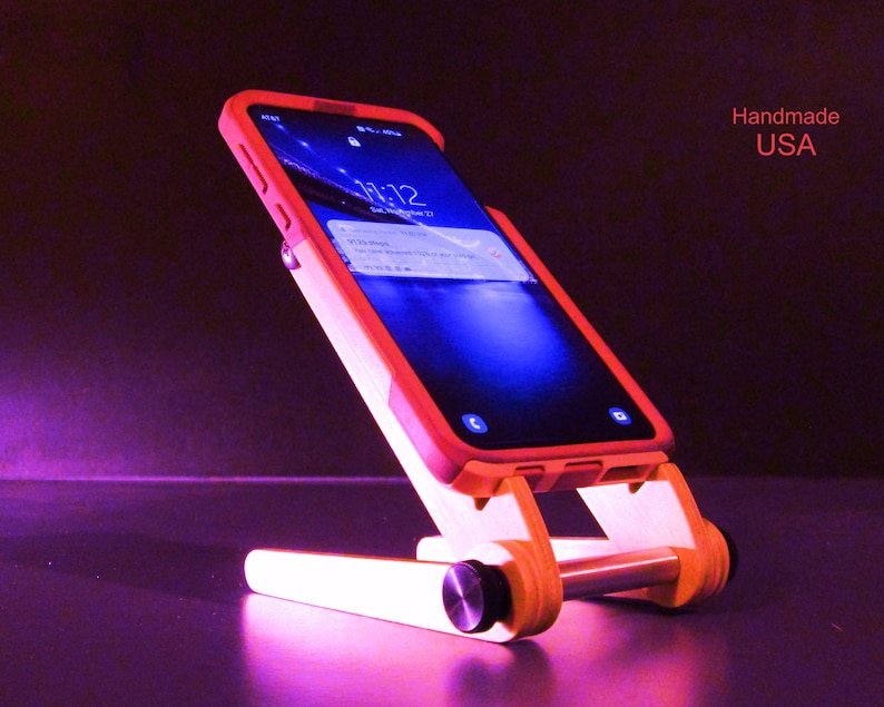 May include: A wooden phone stand with a red-orange phone case. The stand is made of light-colored wood and has a minimalist design. The phone displays the time and date. Text on the image reads "Handmade USA".