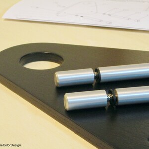 May include: Close-up of a black wooden furniture component with a large circular hole and two silver metal rods. The rods have a cylindrical shape with a rounded end. A sheet of instructions is visible in the background.