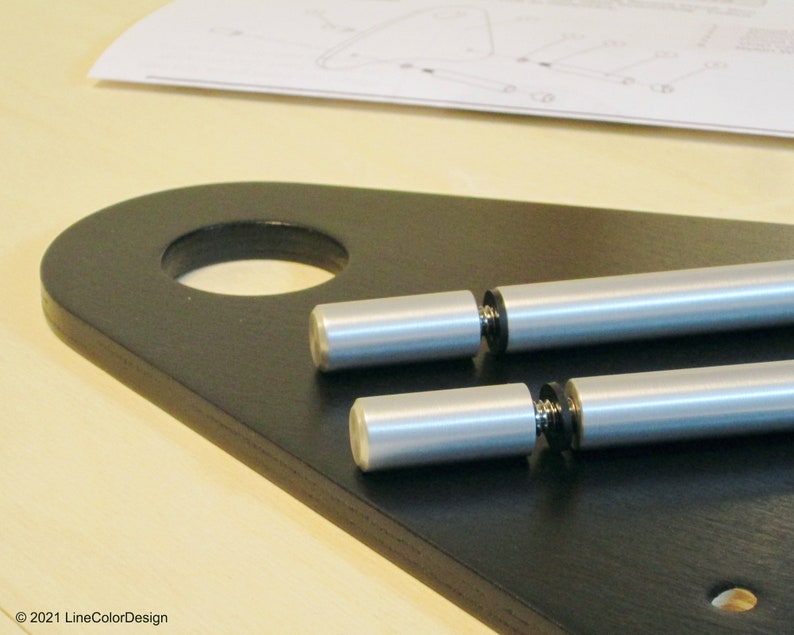 May include: Close-up of a black wooden furniture component with a large circular hole and two silver metal rods. The rods have a cylindrical shape with a rounded end. A sheet of instructions is visible in the background.