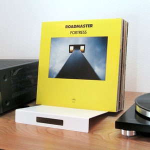 May include: A white vinyl record holder with a rectangular cutout, holding a stack of vinyl records. The album cover is yellow with the text "ROADMASTER FORTRESS" and a photograph of a tall structure. A black turntable and stereo receiver are also visible.