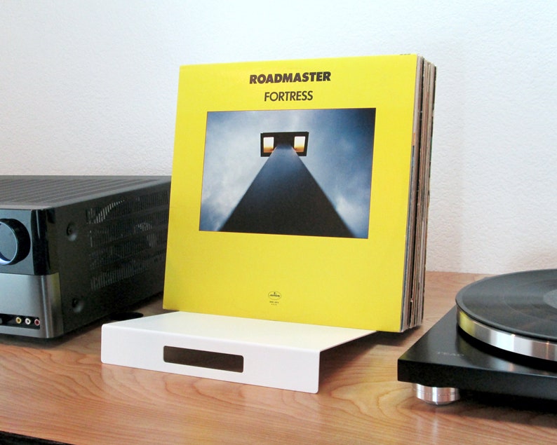 May include: A white vinyl record holder with a rectangular cutout, holding a stack of vinyl records. The album cover is yellow with the text "ROADMASTER FORTRESS" and a photograph of a tall structure. A black turntable and stereo receiver are also visible.