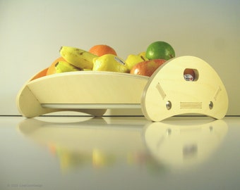 May include: A wooden fruit bowl filled with various fruits, including bananas, oranges, lemons, and apples. A separate wooden serving tray with a dice in the center is next to the bowl. The bowl and tray are light-colored wood.