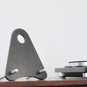 May include: A dark gray, triangular audio equipment stand with a circular hole at the top and two silver pegs. The stand is positioned next to a turntable on a wooden surface, against a white wall.