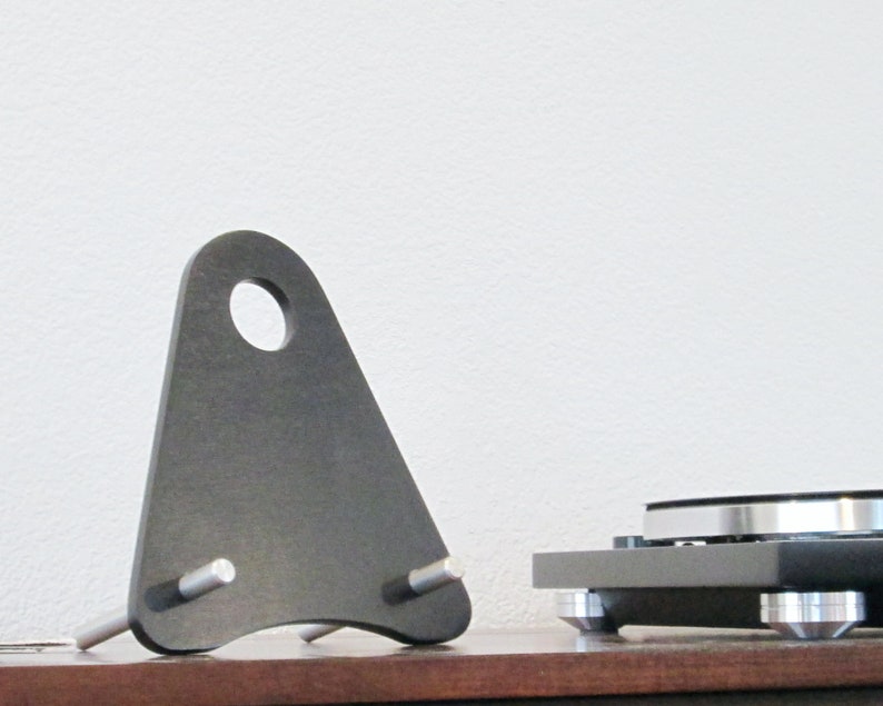 May include: A dark gray, triangular audio equipment stand with a circular hole at the top and two silver pegs. The stand is positioned next to a turntable on a wooden surface, against a white wall.