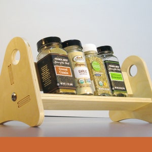 May include: A wooden spice rack with four glass jars of spices. The rack is made of light-colored wood and has a simple, modern design. The jars contain ground cumin, garlic granules, oregano, and rosemary.