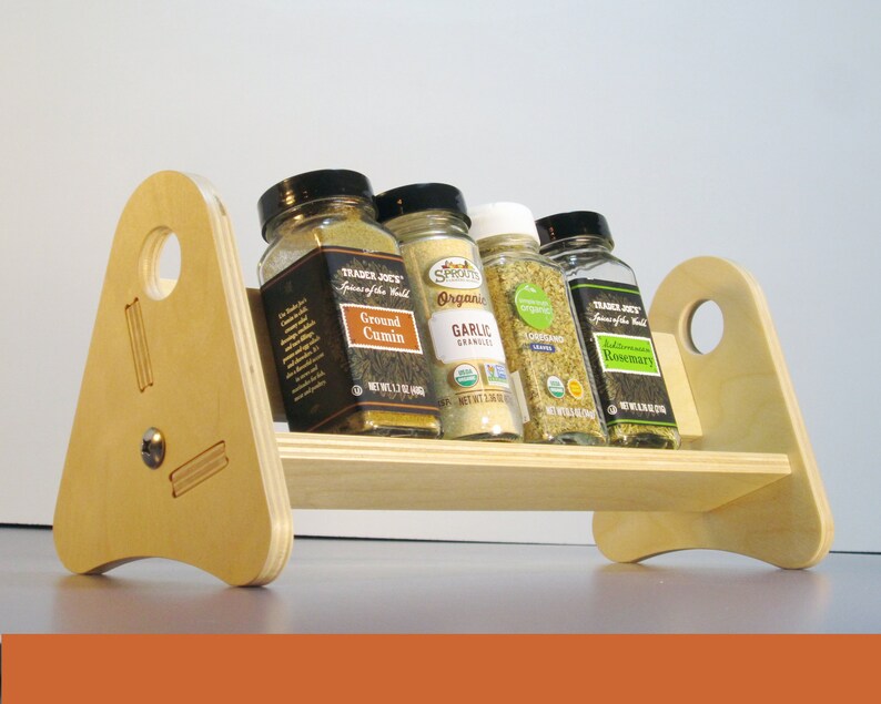 May include: A wooden spice rack with four glass jars of spices. The rack is made of light-colored wood and has a simple, modern design. The jars contain ground cumin, garlic granules, oregano, and rosemary.